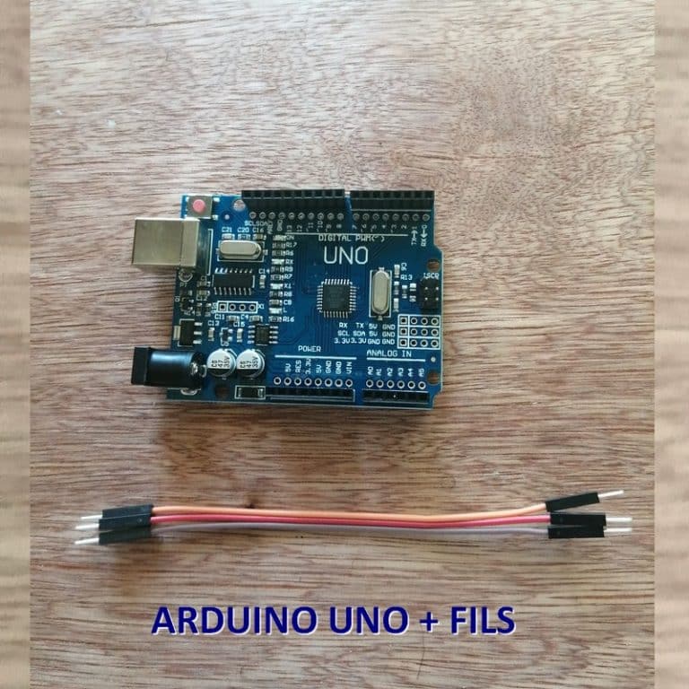 Photo d'une carte Arduino Uno posée sur une surface en bois. L'Arduino est clairement visible, avec tous ses composants et étiquetages détaillés, tels que les ports numériques, les ports d'alimentation et la puce microcontrôleur. Juste en dessous de la carte, il y a trois fils électriques avec des connecteurs aux extrémités, colorés en rouge, noir, et orange, disposés horizontalement. Le texte 'ARDUINO UNO + FILS' est affiché en bas pour identifier le contenu de l'image.