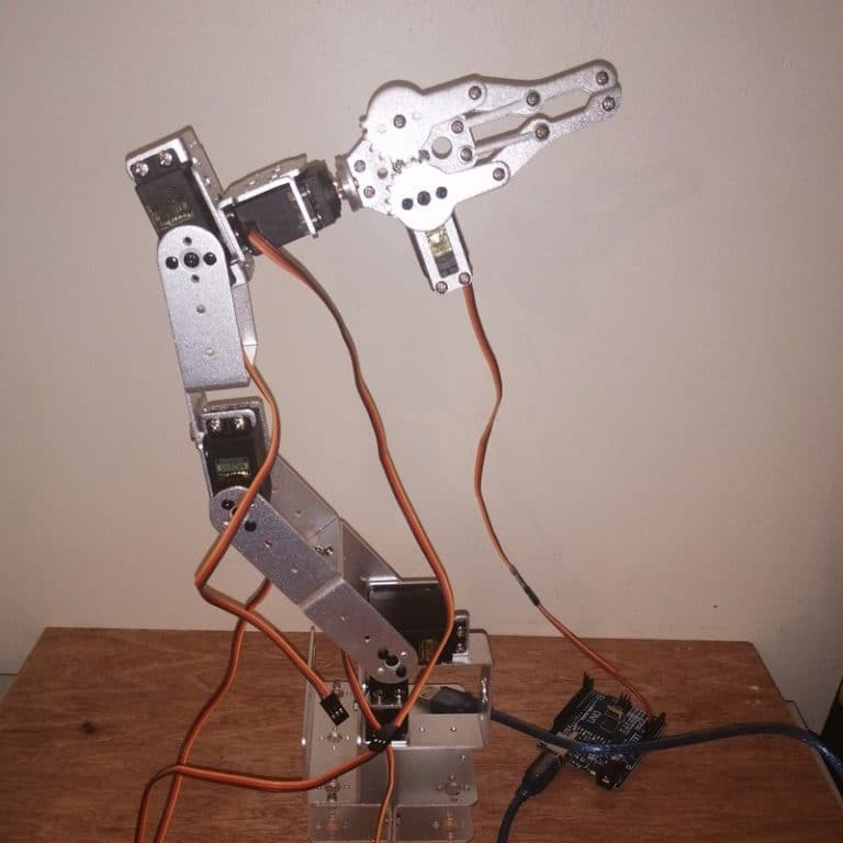Bras robotique articulé en position élevée, connecté à une carte Arduino via des câbles oranges. Le robot est composé de plusieurs segments métalliques avec des servomoteurs visibles, soutenant une pince mécanique complexe en position ouverte. L'ensemble est posé sur un bureau en bois avec un mur uni en arrière-plan