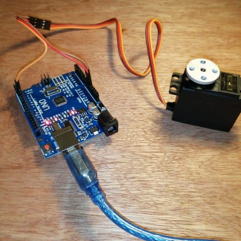 Présente un Arduino Uno connecté à un servomoteur. L'Arduino, alimenté par un câble USB bleu, est utilisé pour contrôler le servomoteur, qui est équipé du palonnier en métal montré dans la première image. Cette configuration illustre comment le palonnier est monté sur le servomoteur.